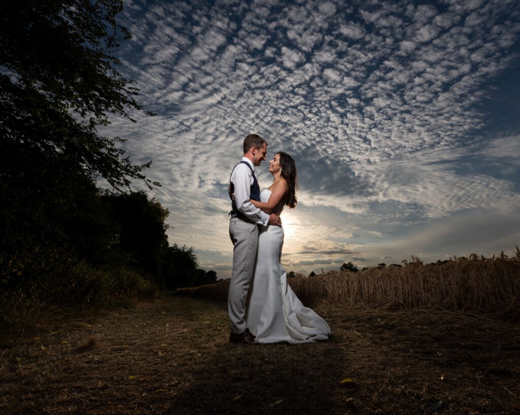 Couple standing together on a countryside path at East Mersea Hall, framed by tall grasses and dramatic sunset clouds, creating a romantic and atmospheric wedding portrait.