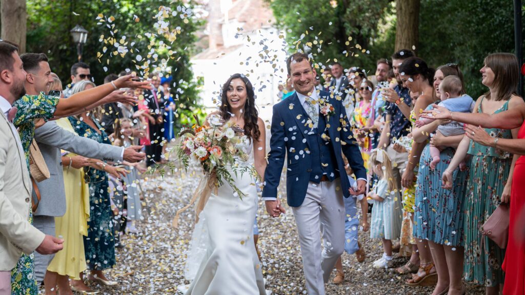 Couple walking hand in hand along the garden path at East Mersea Hall while guests throw confetti, surrounded by greenery and joyful celebration.