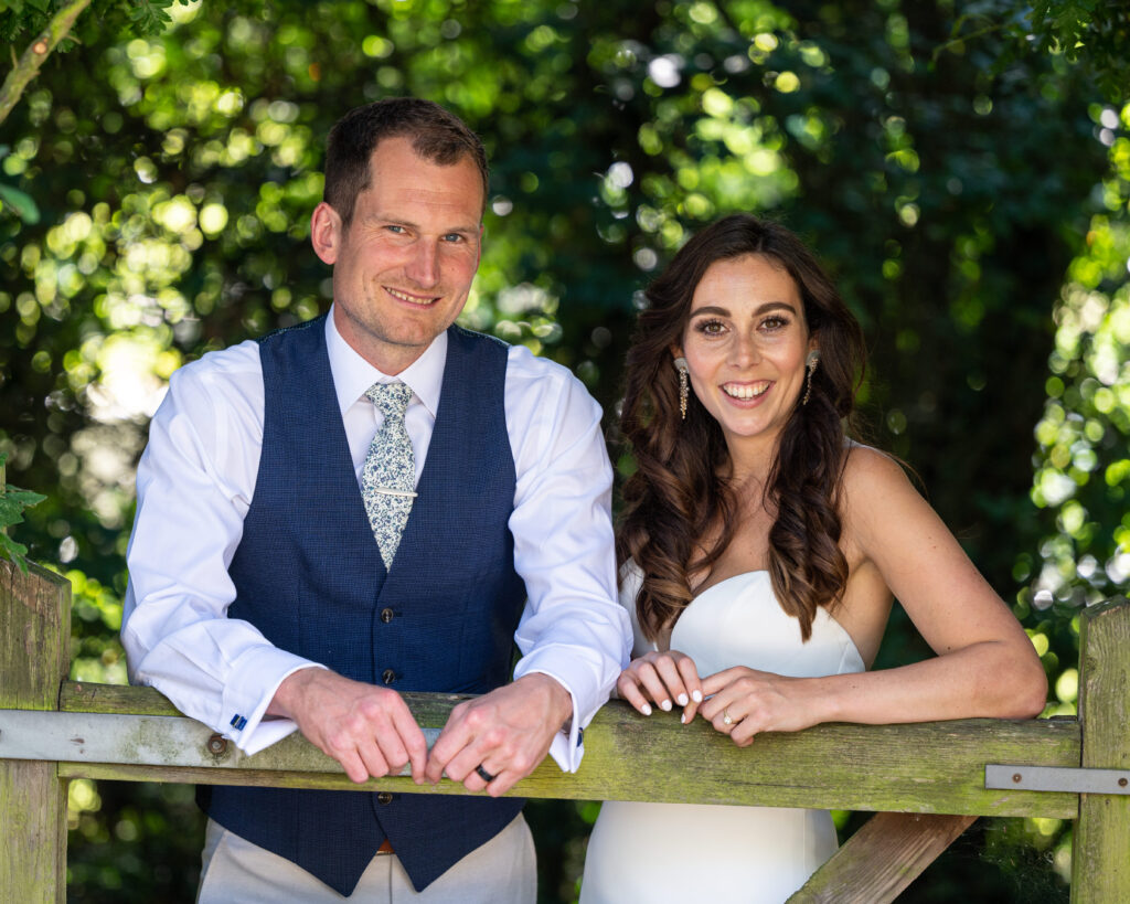 Couple standing behind a wooden gate at East Mersea Hall, surrounded by lush greenery and soft sunlight, creating a relaxed and intimate wedding portrait.