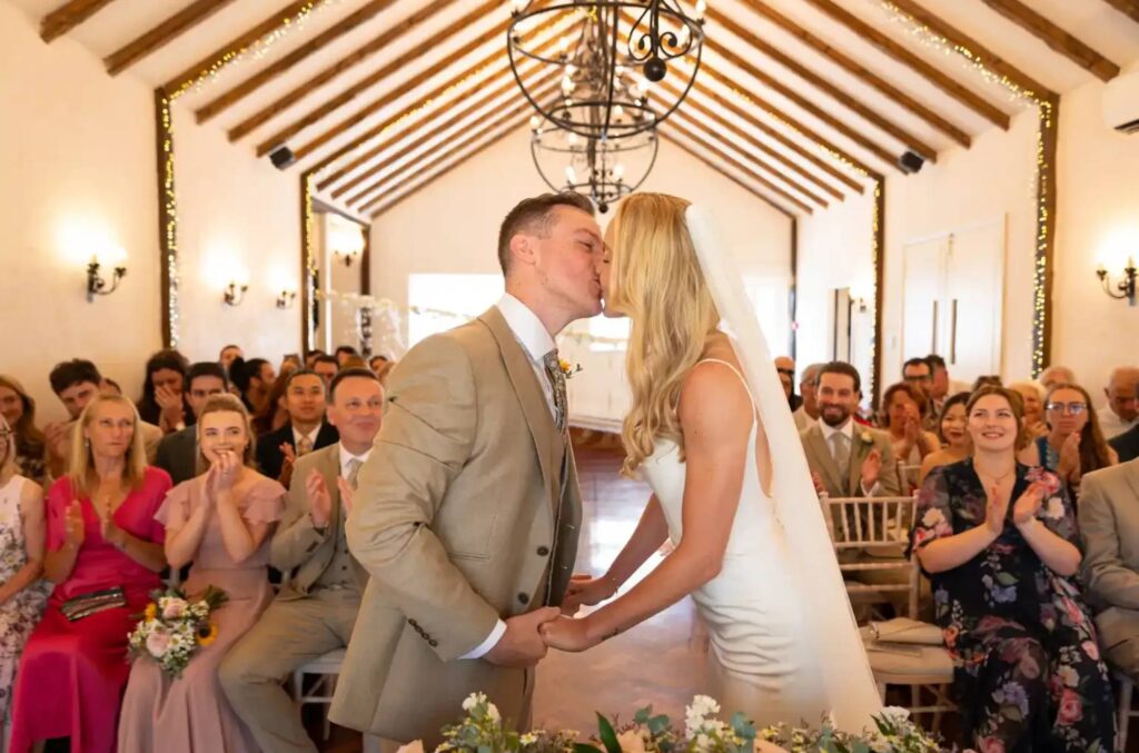 Newly married couple sharing a joyful kiss during their ceremony, as guests applaud and the officiant looks on.
