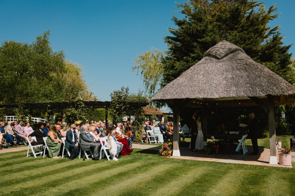 Outdoor wedding ceremony at a thatched‑roof gazebo, with the couple and celebrant surrounded by garden greenery and seated guests on the lawn.