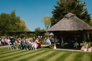 Outdoor wedding ceremony at a thatched‑roof gazebo, with the couple and celebrant surrounded by garden greenery and seated guests on the lawn.
