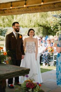 Outdoor wedding ceremony under a wooden structure with string lights, as the couple hold hands and look toward the guest giving a reading, with family and friends seated in the garden.
