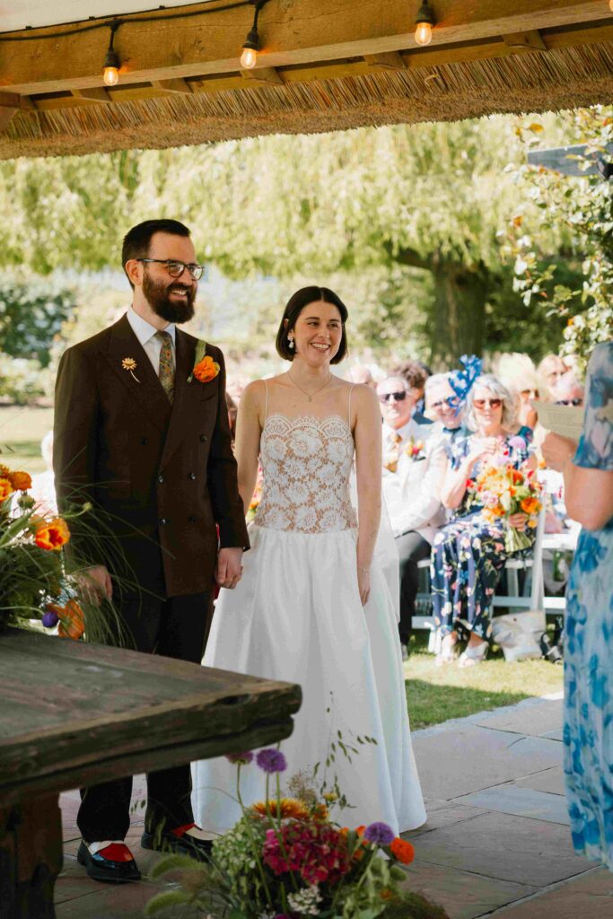 Outdoor wedding ceremony under a wooden structure with string lights, as the couple hold hands and look toward the guest giving a reading, with family and friends seated in the garden.