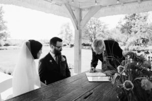 Celebrant signing the wedding certificate as the couple sit together under a wooden gazebo, with flowers on the table and garden greenery behind them.