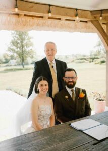 Celebrant standing with the couple under a thatched wooden gazebo, with the open certificate on the table and garden views behind them.