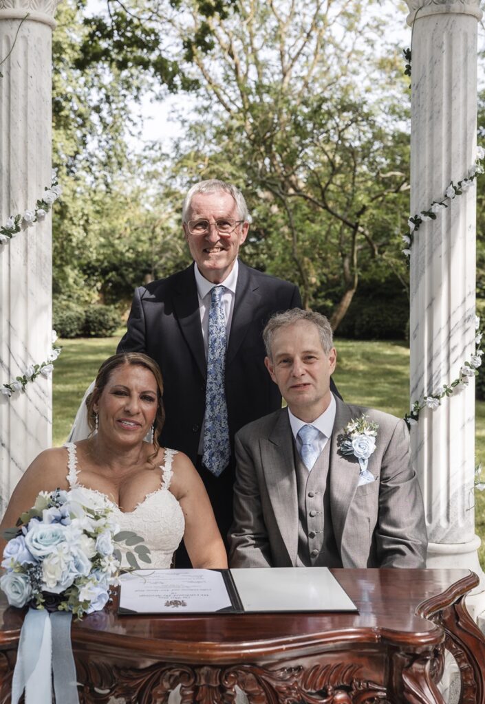 A bride and groom after signing their certificate under a flower‑decorated pergola, with their celebrant standing behind them.