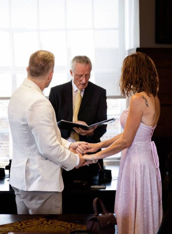 The couple holding hands during their Essex vow renewal as the celebrant reads from a ceremony script.